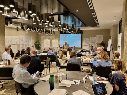 Conference room workshop with participants at round tables; presenter at front near a screen titled 'Discussion: Zoning & Urban Planning Regulations'.